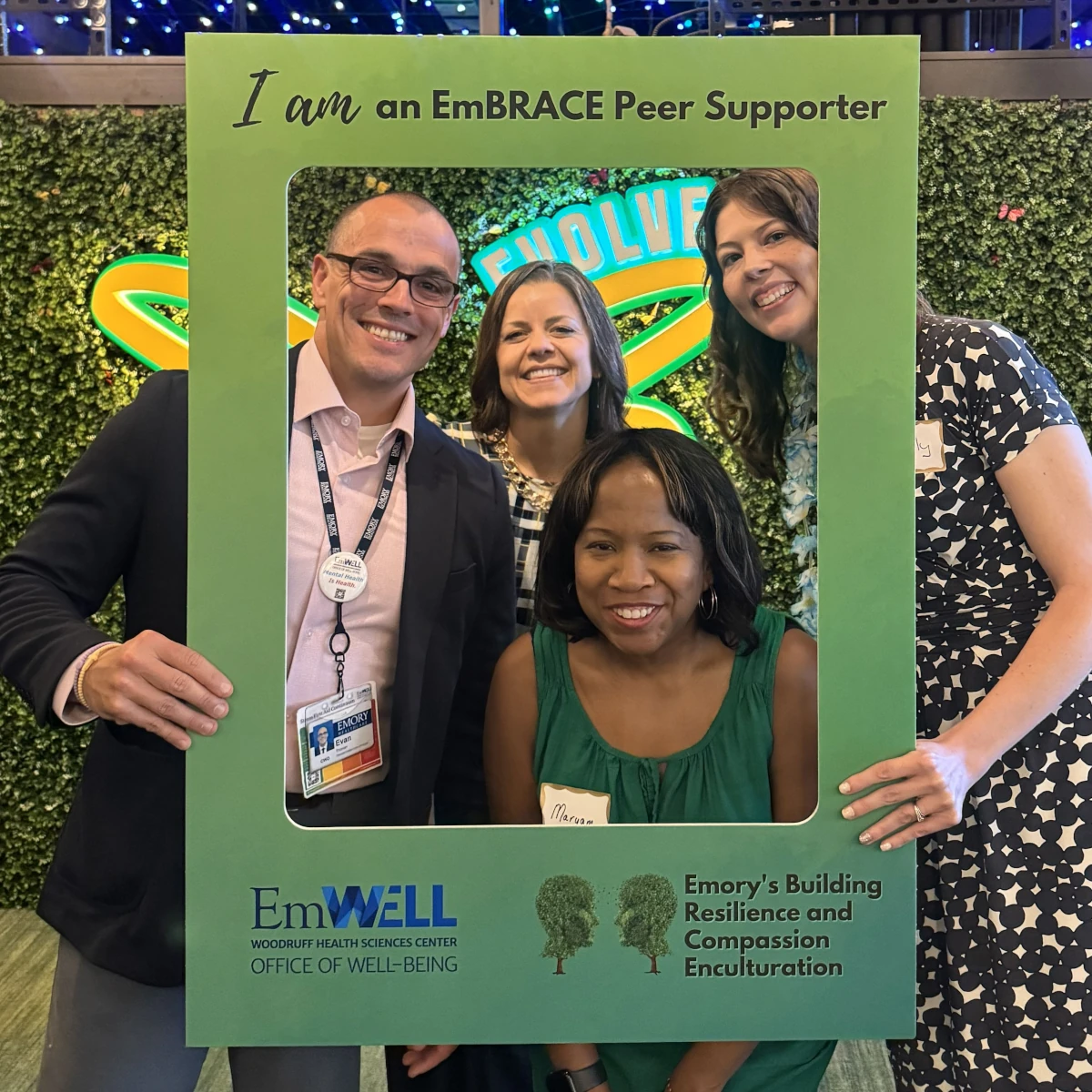 Four Emory employees posing together with a green photo frame that reads 'I am an EmBRACE Peer Supporter' at an EmWELL event