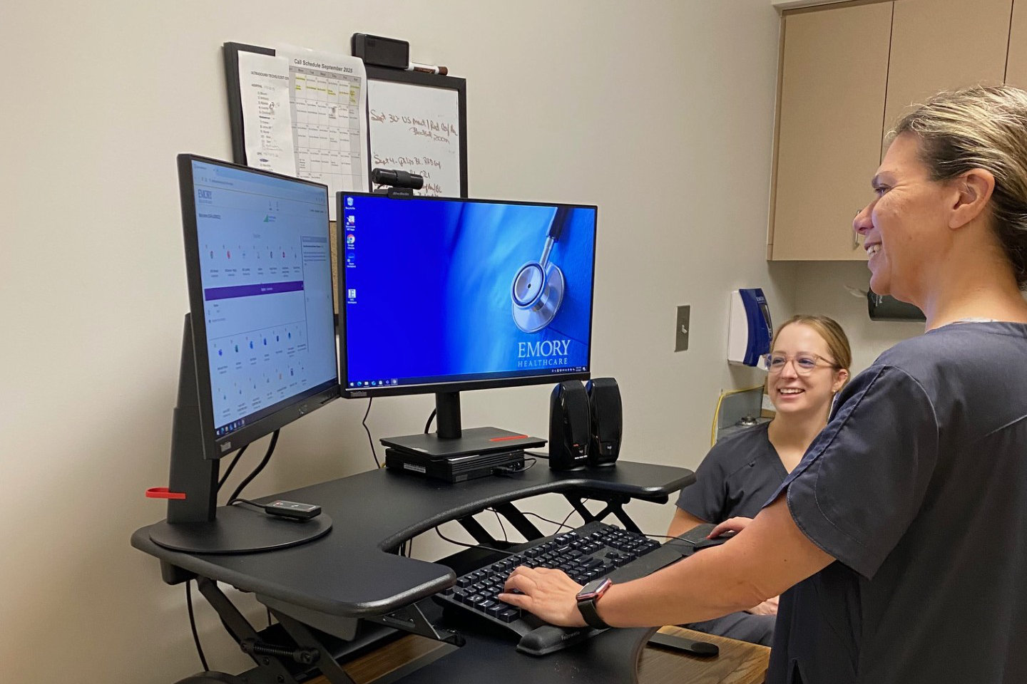 Two Emory Healthcare staff members smiling while working at a standing desk with dual monitors