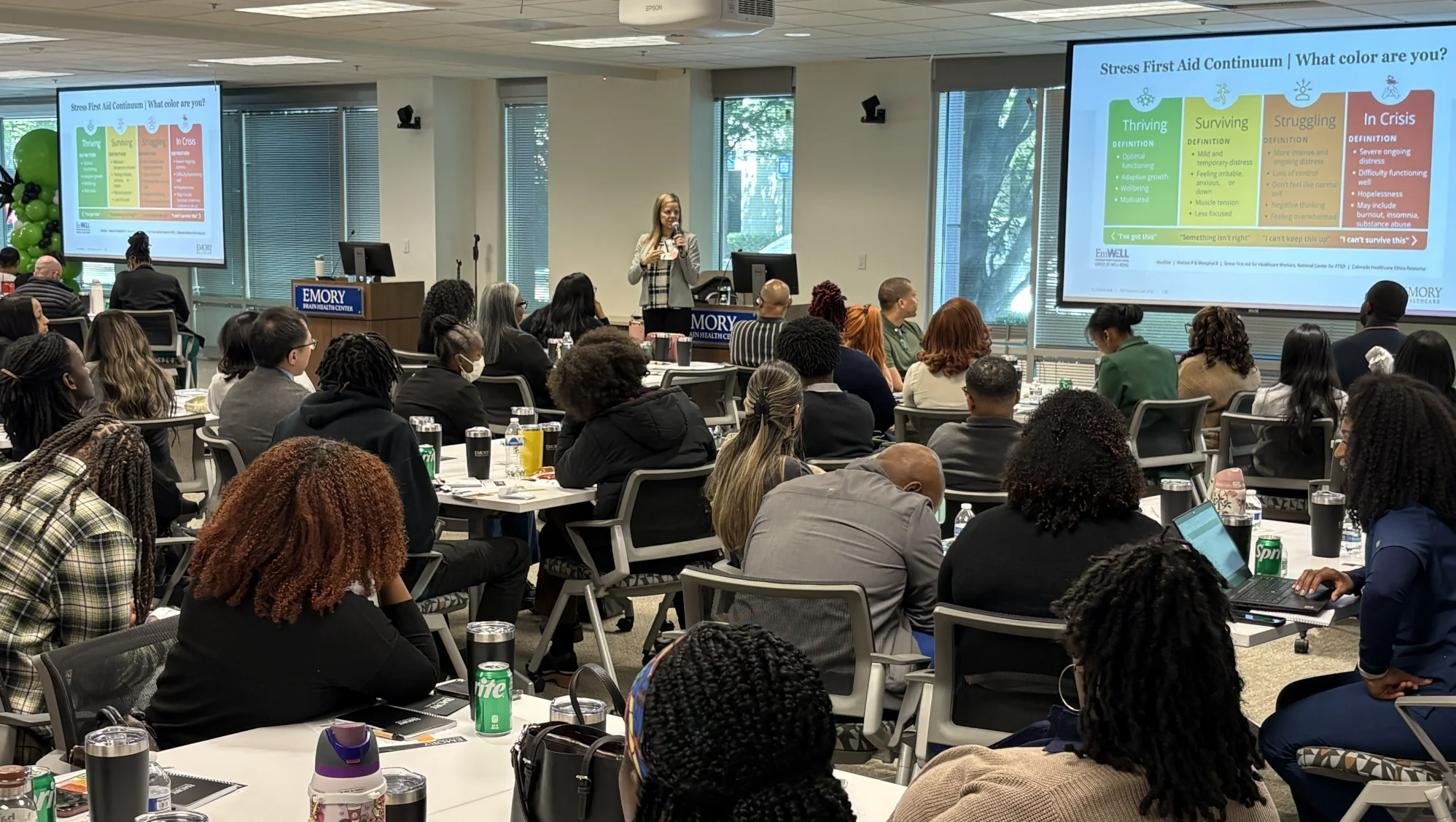 EmWELL Stress First Aid training session at Emory Healthcare with a presenter speaking to a room of several dozen employees. Two projection screens display the Stress First Aid Continuum.