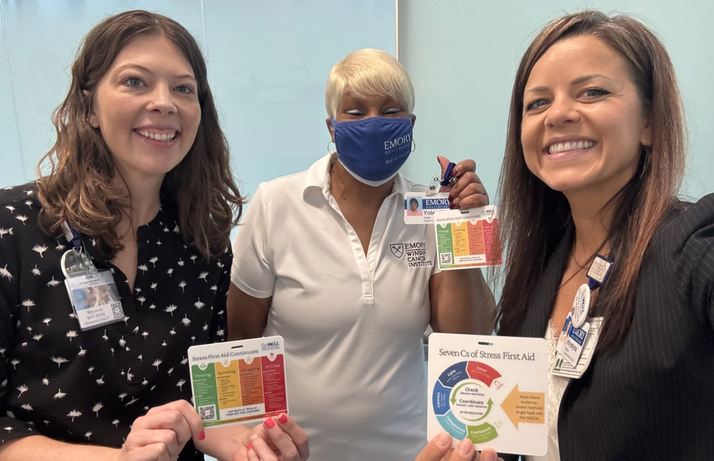 Three Emory Healthcare staff members displaying Stress First Aid resource cards, including the Stress First Aid Continuum and Seven Cs of Stress First Aid.